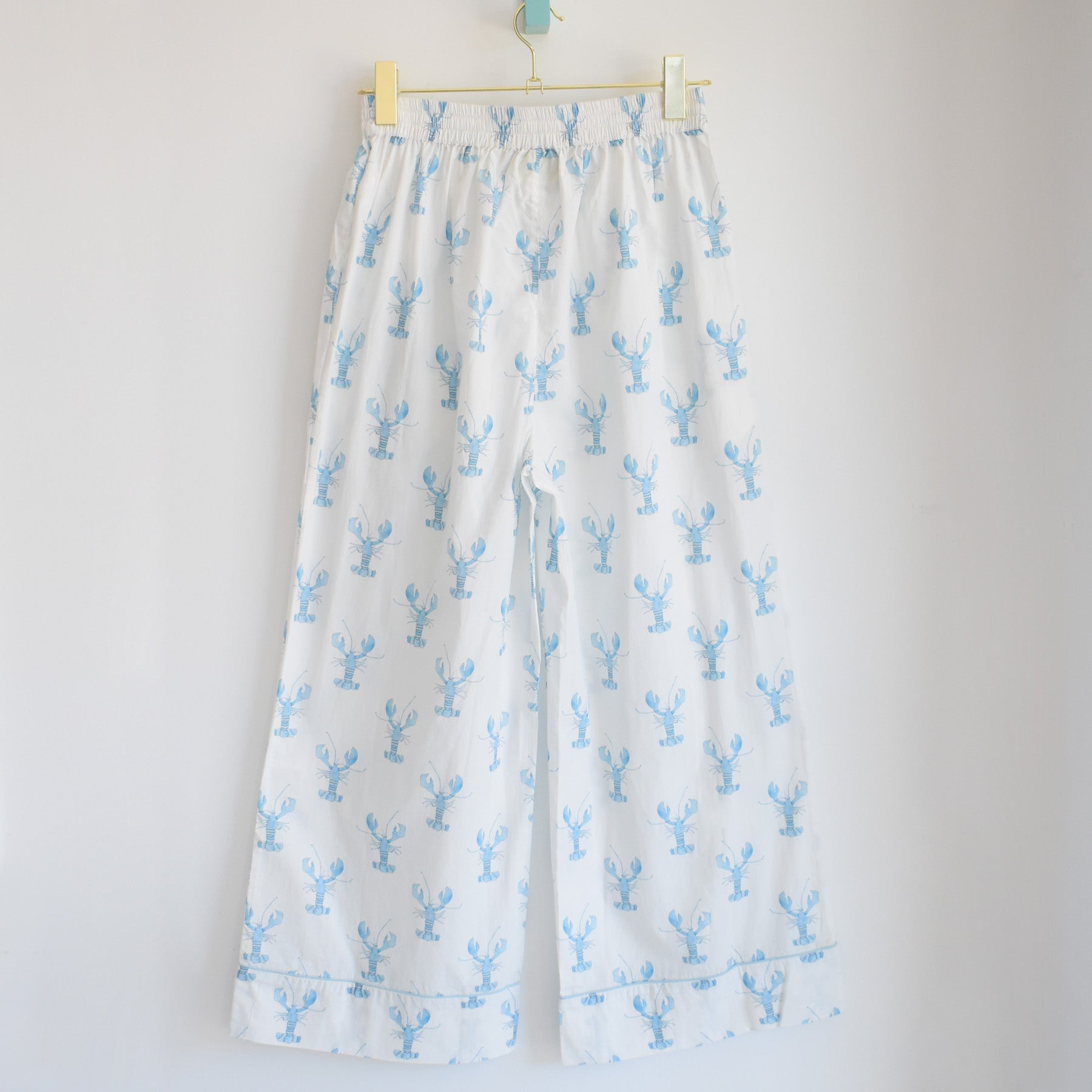 White pajama pants featuring a light blue lobster print hang on a wooden hanger against a plain white wall, showcasing the Sara Fitz Blue Lobster Pants Pajama Set—crafted from 100% cotton and perfect for relaxed coastal-inspired sleepwear.
