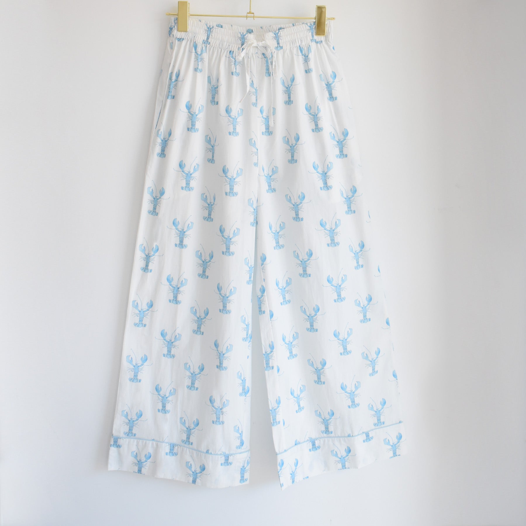 White wide-leg pajama pants featuring a light blue lobster print, from the Sara Fitz Blue Lobster Pants Pajama Set, are shown hanging on a wooden hanger against a plain white background. Made from 100% cotton pajama fabric, these cozy bottoms embody the charm of coastal sleepwear.