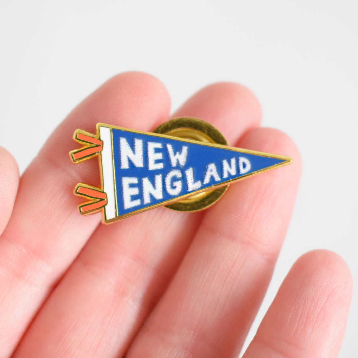 A hand holds the Sara Fitz Pennant Needle Minder, a small, triangular blue and yellow enamel pin shaped like a pennant with "NEW ENGLAND" in white letters, serving as a charming accessory for your stitching supplies.