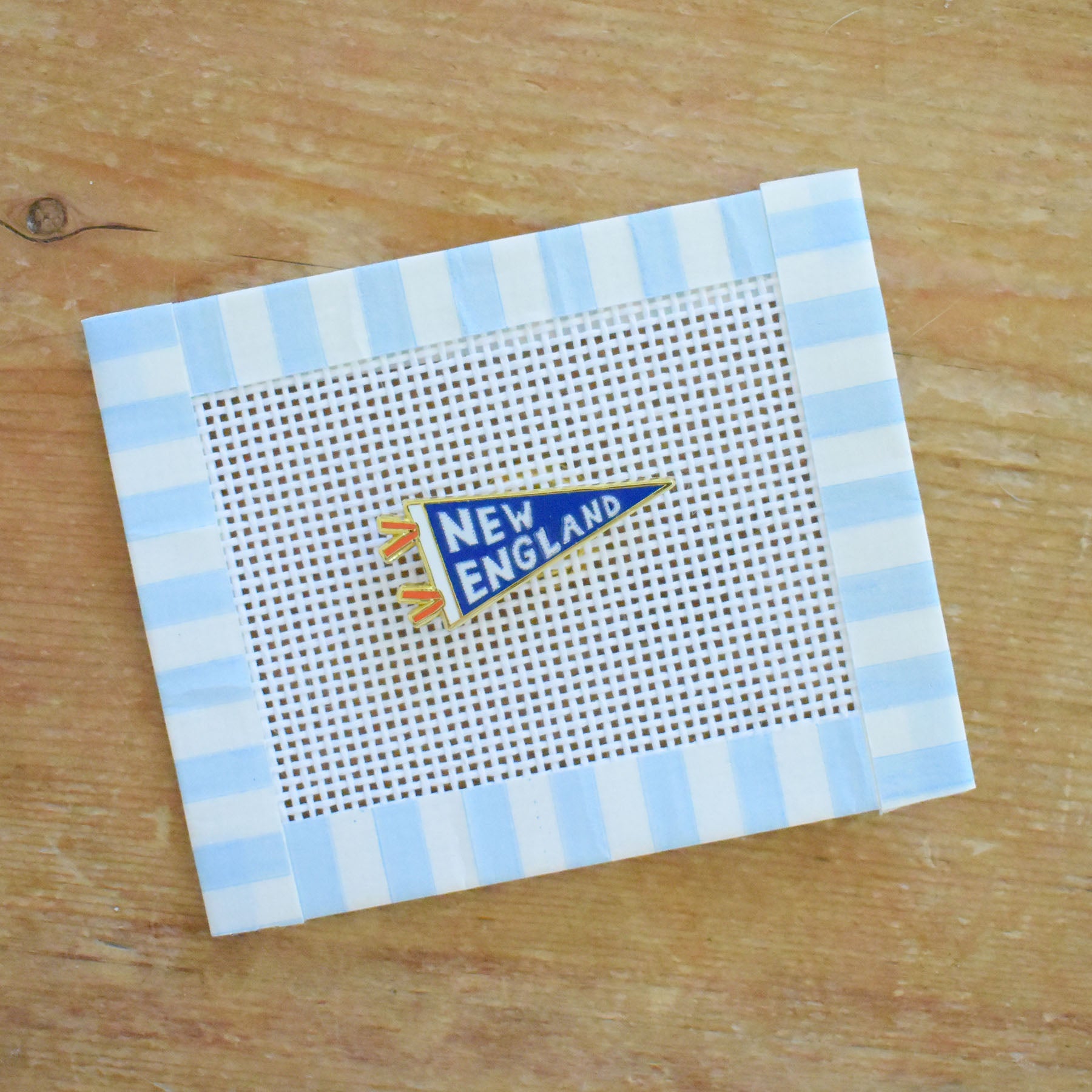 A small needlepoint canvas featuring a New England pennant design, bordered with blue and white striped tape, sits atop a wooden surface. Next to it is the Sara Fitz Pennant Needle Minder—an ideal stitching accessory that keeps your needle secure while you work.