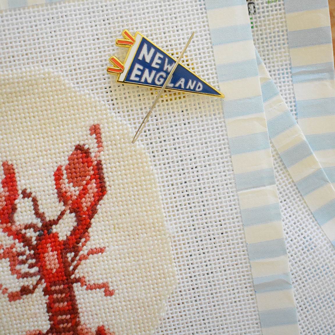 A needlepoint canvas featuring a red lobster design, a needle, and Sara Fitz’s blue New England pennant pin, all arranged atop light blue and white striped fabric—perfectly complemented by the Pennant Needle Minder from Sara Fitz as your essential stitching accessory.