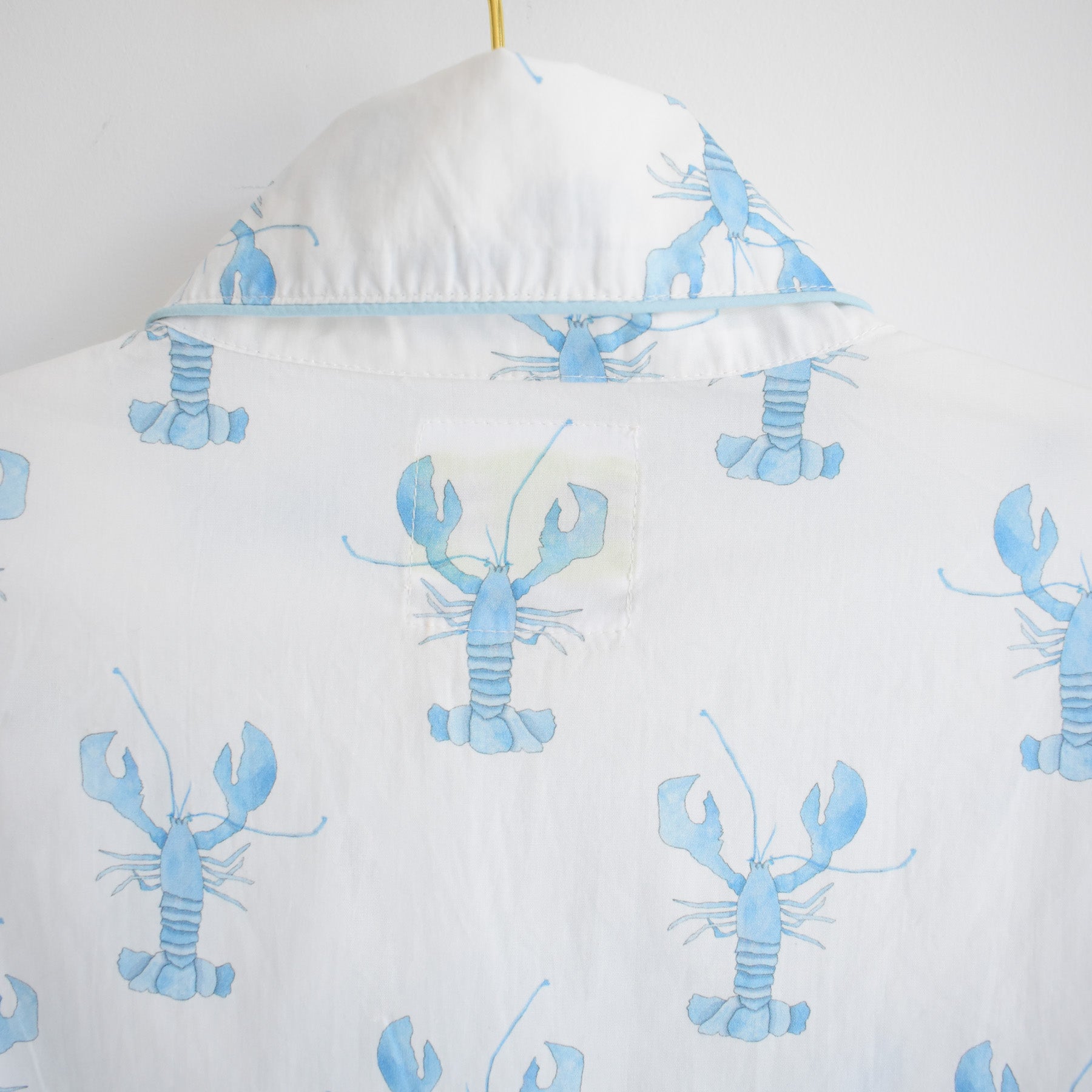 A back view of the Sara Fitz Blue Lobster Short Pajama Set showcases a crisp white shirt adorned with a light blue lobster pattern. The collar is visible, and one lobster near the neckline appears slightly faded or patchy, enhancing the relaxed charm of this cotton pajama set—an ideal choice for anyone seeking cozy coastal pajamas.