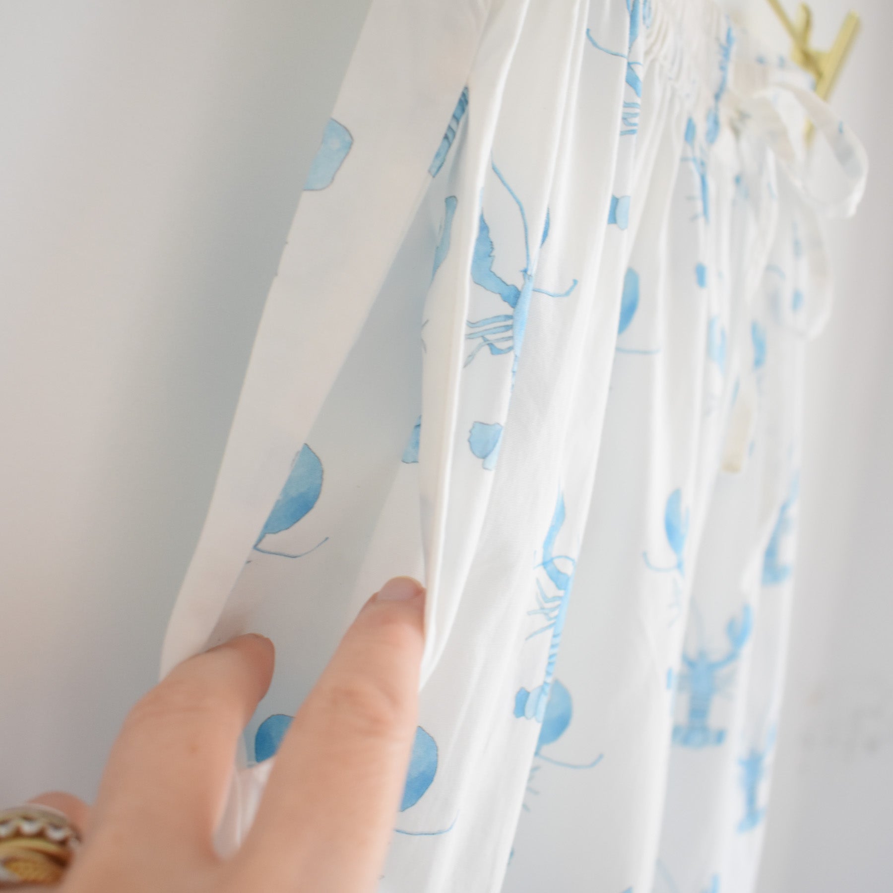 A hand holds the side seam of a white dress featuring light blue lobster prints, revealing a hidden pocket—an adorable detail inspired by our Sara Fitz Blue Lobster Short Pajama Set.