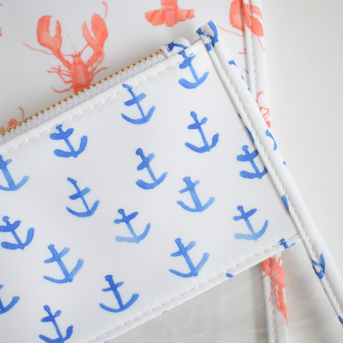The Anchor Project Bag by Sara Fitz—a white pouch adorned with blue anchor prints and made from heavyweight canvas—rests atop fabric decorated with orange lobster illustrations. Both pieces share a nautical theme inspired by Sara Fitz’s signature style, and the bag’s gold zipper stands out.