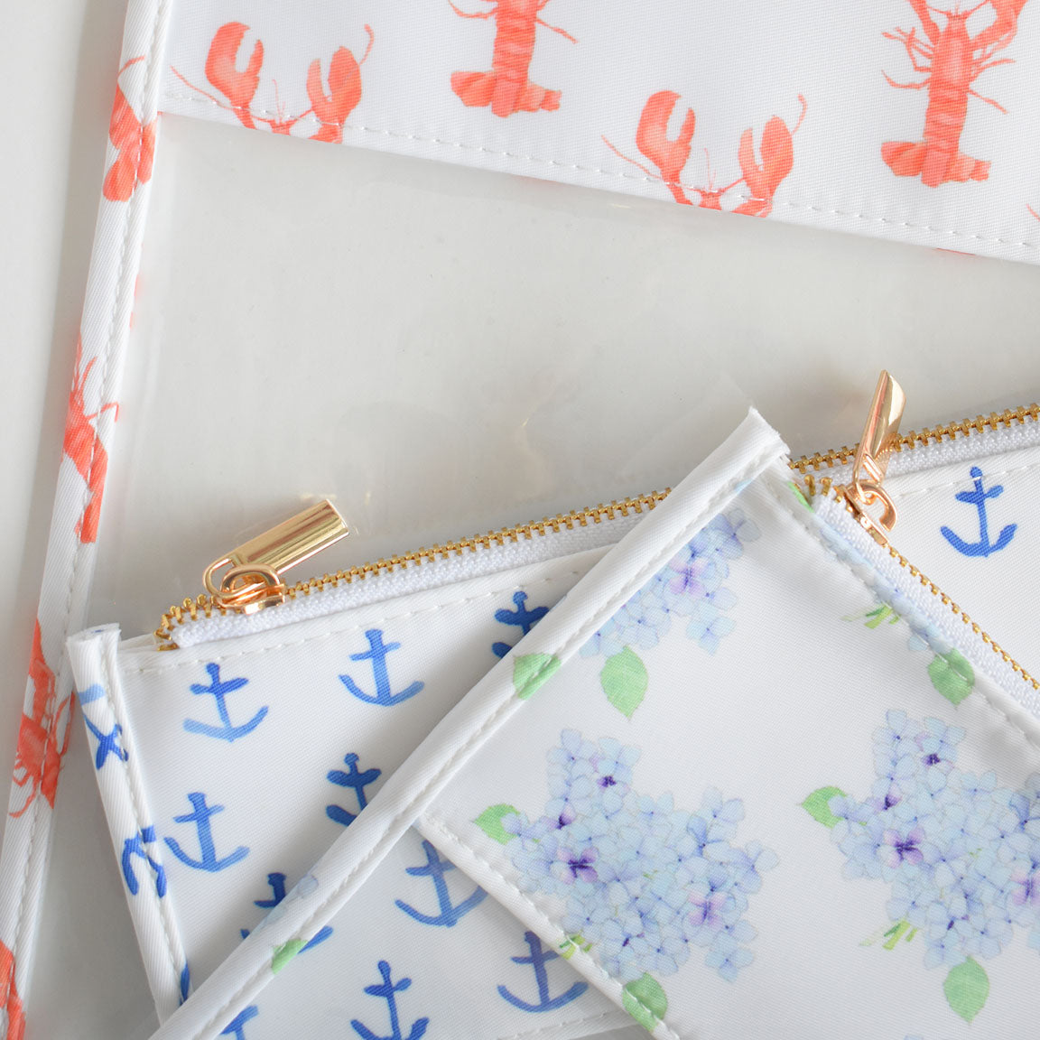 Three Sara Fitz project bags overlap. One showcases red lobsters, another displays blue anchors, and the third features light blue and purple hydrangeas with green leaves. All are made from heavyweight canvas with gold zippers, arranged beautifully on a white surface.