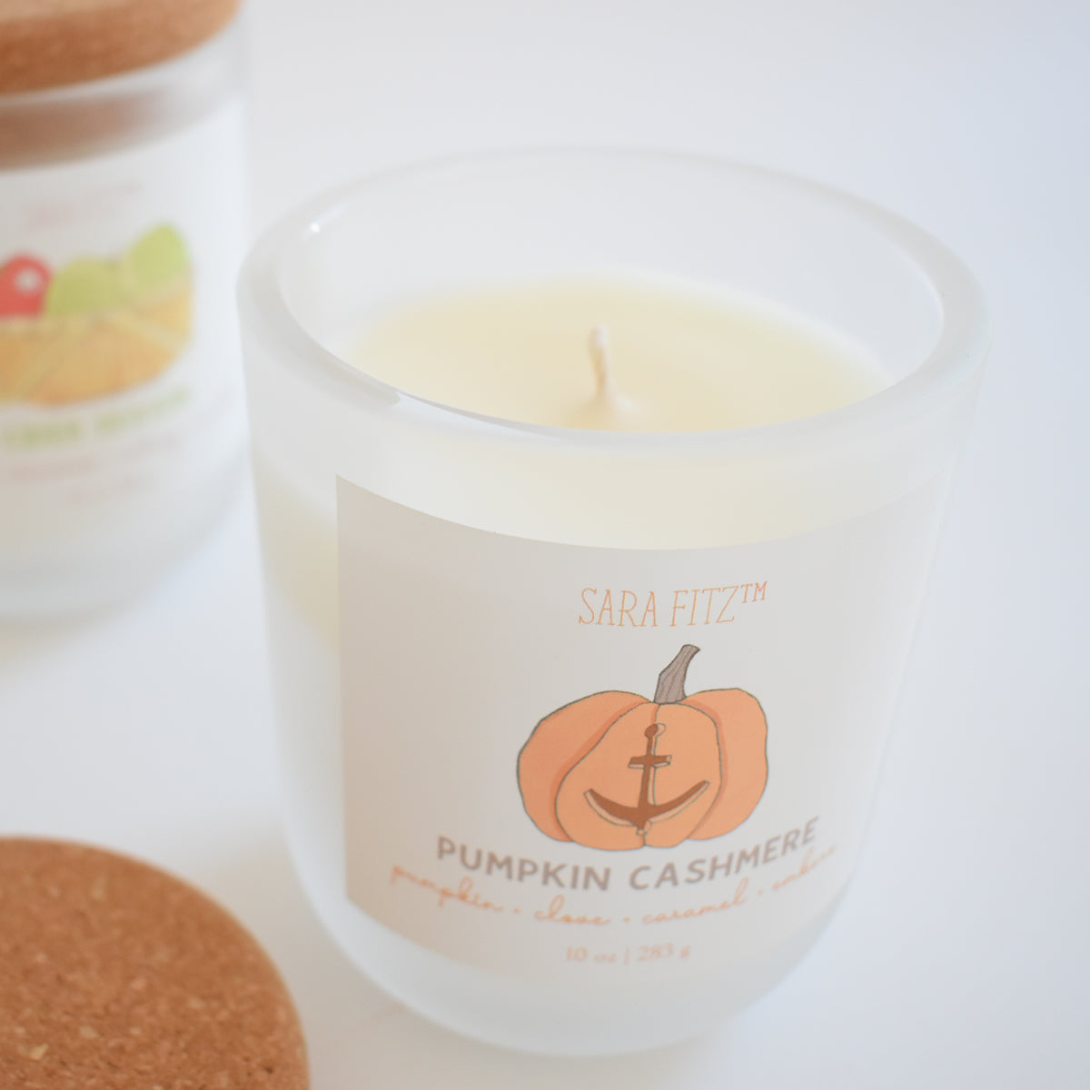 A white frosted glass Pumpkin Cashmere Candle by Sara Fitz, featuring an orange pumpkin and anchor design, is hand-poured in Maine. The candle has a cork lid positioned nearby, while another similar candle appears blurred in the background.