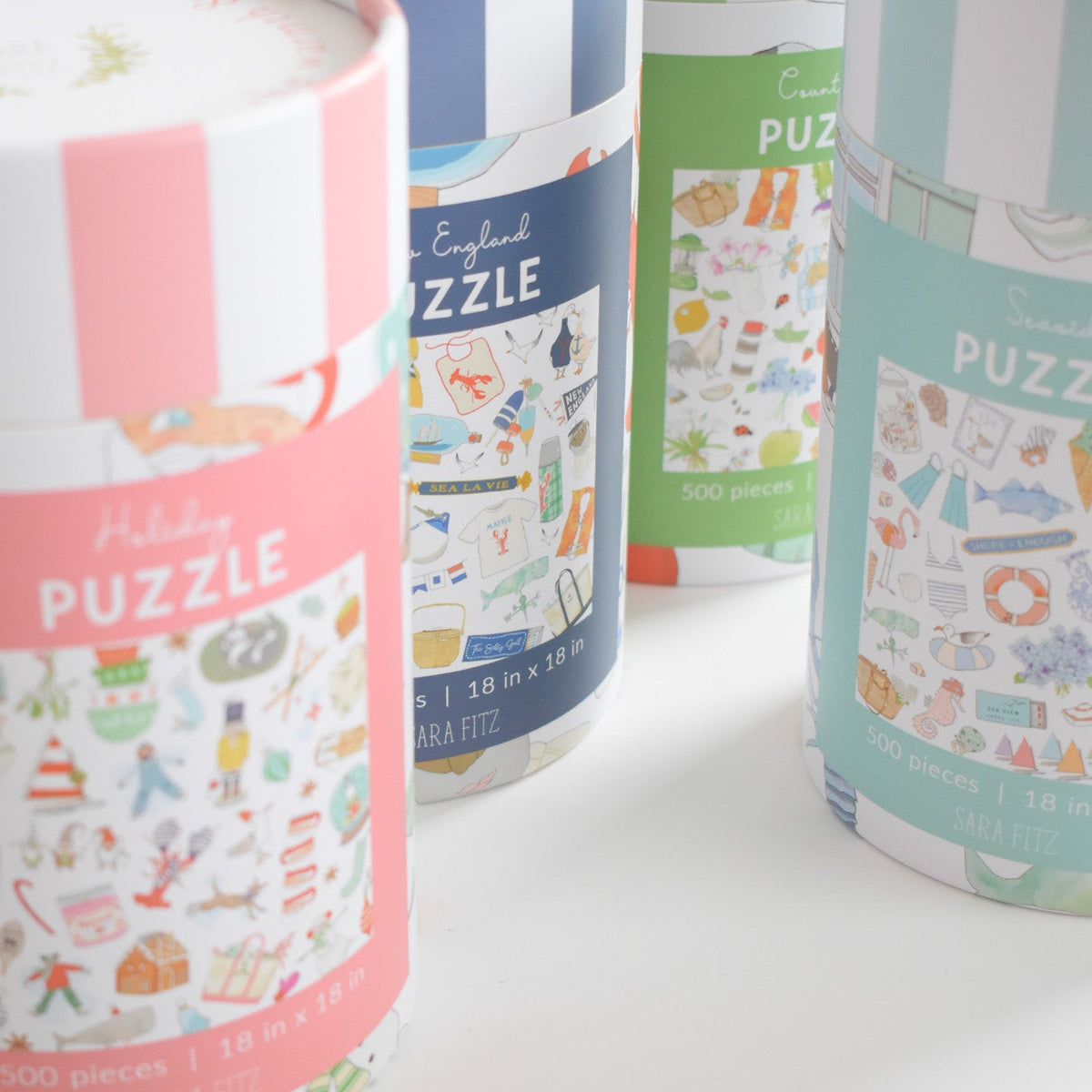 Four cylindrical 500-piece jigsaw puzzle containers with watercolor illustrations, including a pink Holiday puzzle, a blue New England puzzle, and a light blue Seaside Puzzle.