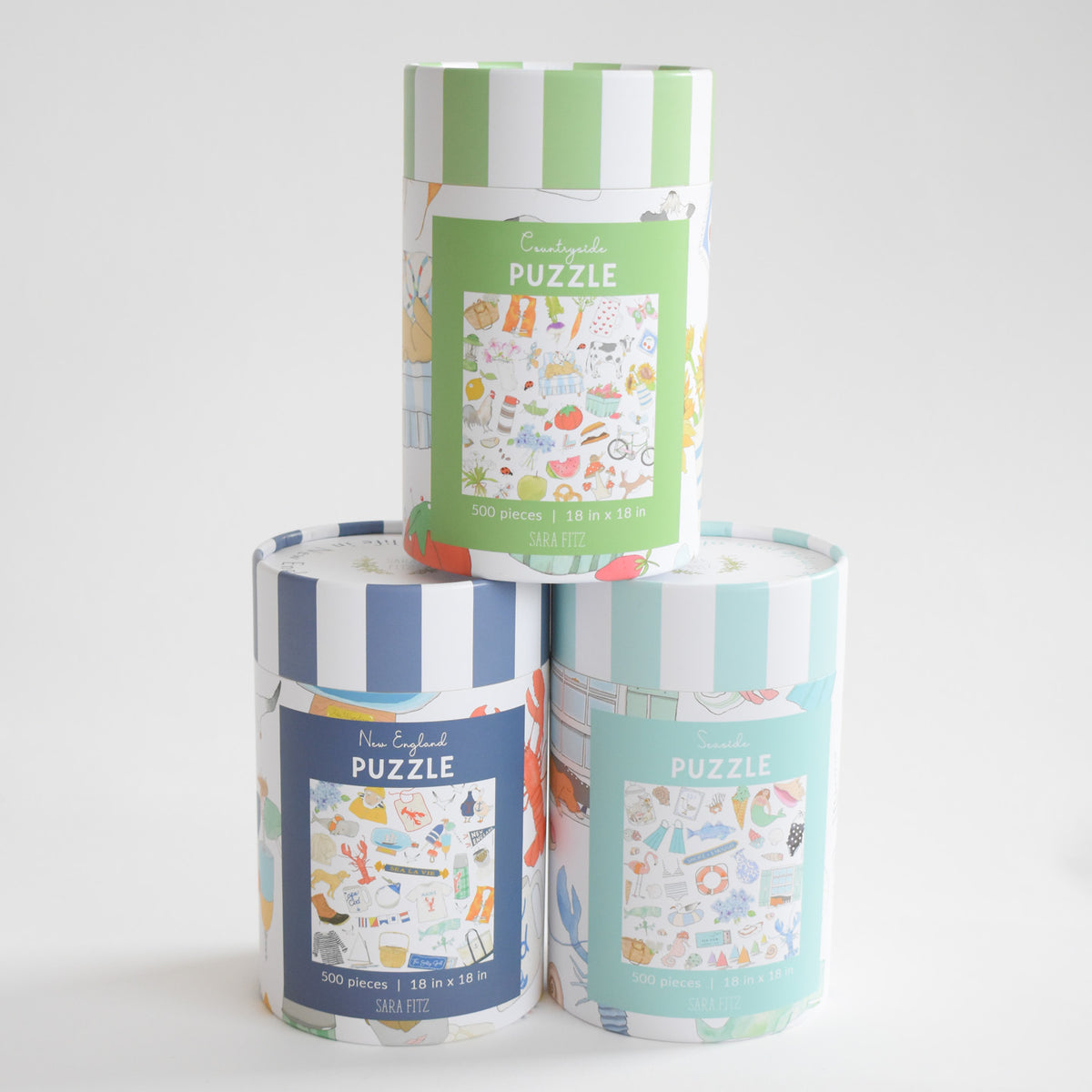 Three cylindrical 500-piece Seaside Puzzle boxes stacked in a pyramid, each featuring colorful illustrations of various seaside or countryside items, including a beach collage.