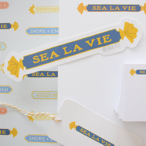 The Sara Fitz Quarterboard Sticker features “SEA LA VIE” in yellow on a blue banner with yellow shell accents, alongside nautical-themed tags like DRIFTWOOD and GULLS NEST. Weatherproof vinyl. Made in the USA.