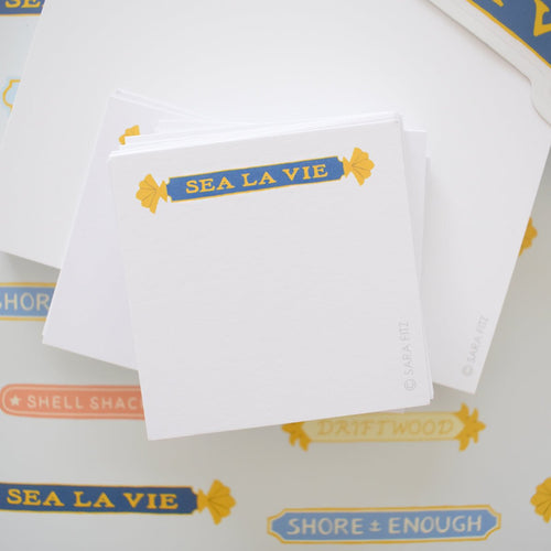 The Sara Fitz Quarterboard Notelet features a stack of white single-sided notelets topped with a blue and yellow "SEA LA VIE" banner, surrounded by ocean-themed stickers, all printed in the USA.