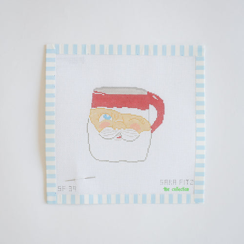 The Santa Mug Needlepoint by Sara Fitz features a cross-stitch Santa Claus mug on white 18-mesh canvas with blue and white striped borders, red hat and handle, plus “SARA FITZ” stitched at the bottom right.