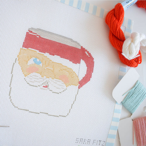 The Sara Fitz Santa Mug Needlepoint features a partially completed mug with a red handle and rim, blue eye, blush details on 18 mesh canvas. Colorful embroidery threads and bobbins surround the project.