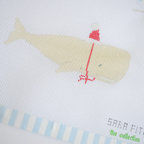 The Santa Whale Needlepoint by Sara Fitz features a light yellow whale in a red Santa hat and scarf on white fabric, with partial “SARA FIT...” text. Ideal for 18 mesh or needlepoint canvas projects.