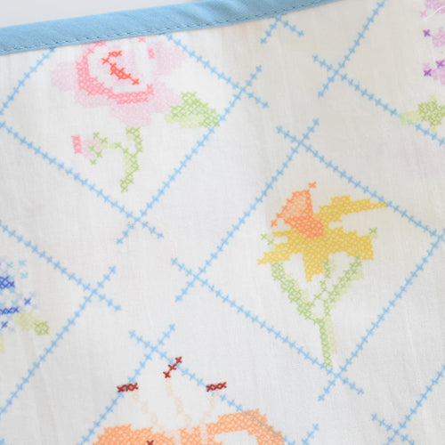 The Sara Fitz Dog Bandana, Cross Stitch Floral, features pastel floral patterns in a blue diamond grid with a blue border—a charming cotton accessory perfect for rescue pups.