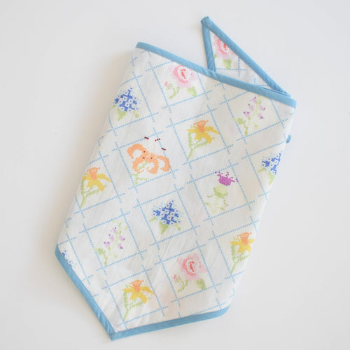The Sara Fitz Dog Bandana, Cross Stitch Floral, is a white quilted cotton bandana with a blue border, diamond pattern, and colorful floral designs, shown folded on a white surface.