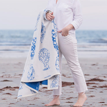 A person in white clothes stands on a sandy beach, holding a white blanket with blue vase patterns. The ocean and sky are visible in the background.