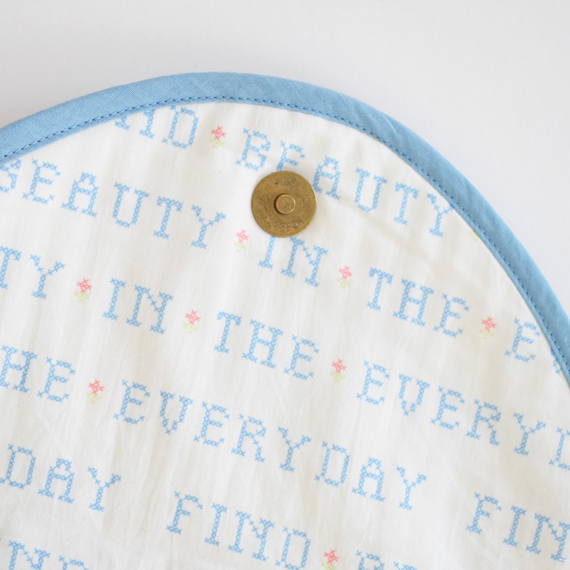 The Sara Fitz Clutch, Cross Stitch Floral, is a white quilted bag with light blue trim, featuring cross-stitched blue "FIND BEAUTY IN THE EVERYDAY" text and a round brass button at the top center.
