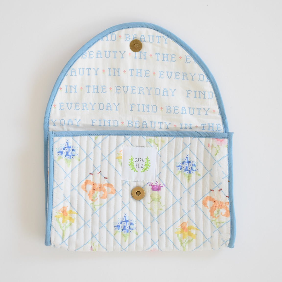 The Sara Fitz Clutch, Cross Stitch Floral features a quilted design with blue border, brass snaps, and floral cross-stitch. The open flap reveals “FIND BEAUTY IN THE EVERYDAY” in pastels and a Sara Fitz label inside this handmade accessory.