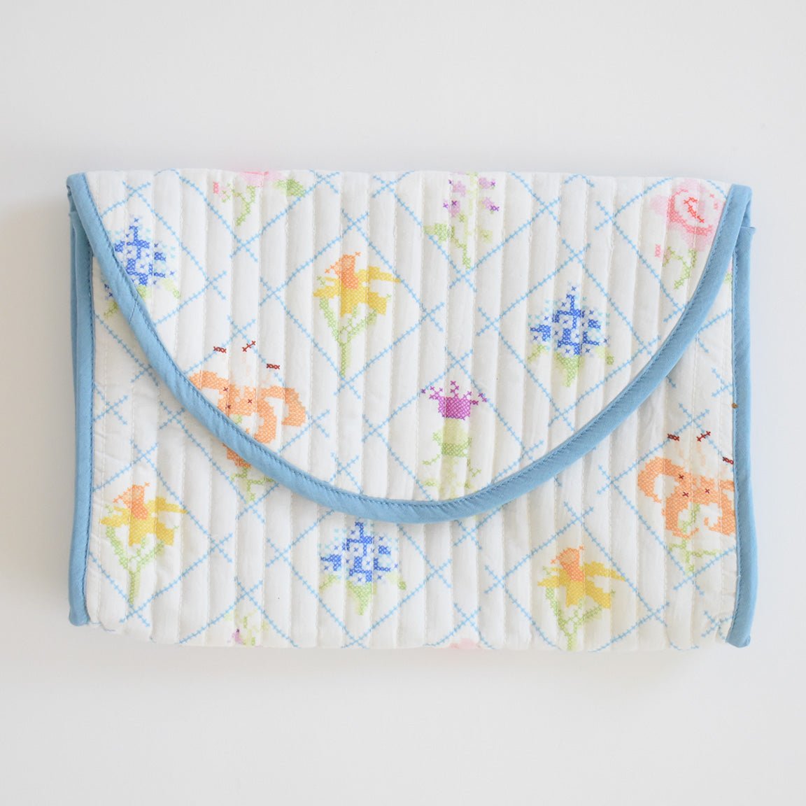 The Sara Fitz Clutch, Cross Stitch Floral, is a handmade quilted cotton clutch with a blue border and pastel floral cross-stitch on white with light blue diamond lines, featuring a fold-over flap for convenience.