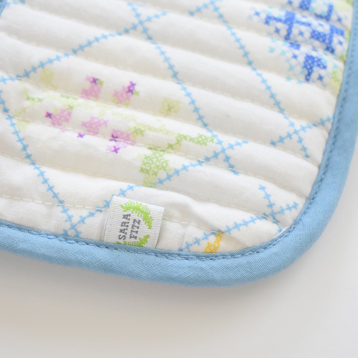A close-up of the Sara Fitz Coin Pouch, Cross Stitch Floral, shows white quilted cotton with blue, green, purple, and yellow floral cross-stitch designs, a light blue border, and a small white tag with green detail near the zip closure.
