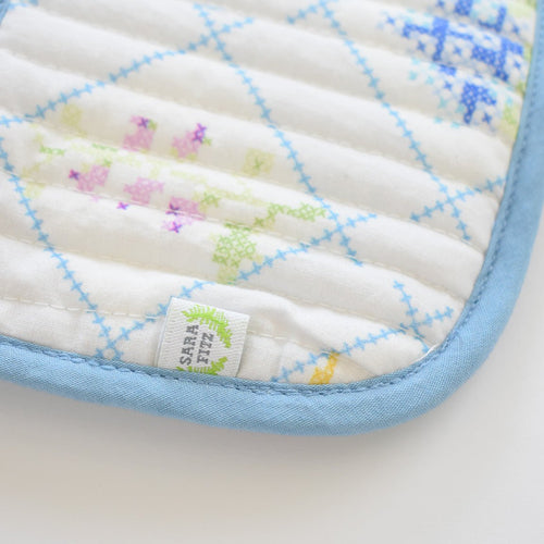 A close-up of the Sara Fitz Coin Pouch, Cross Stitch Floral, shows white quilted cotton with blue, green, purple, and yellow floral cross-stitch designs, a light blue border, and a small white tag with green detail near the zip closure.