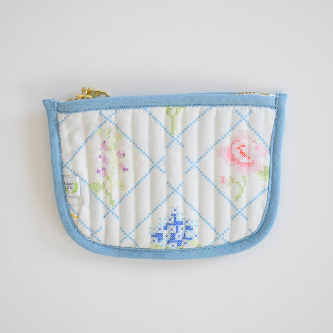 The Sara Fitz Coin Pouch, Cross Stitch Floral, is a quilted cotton card holder with a light blue border and zip closure, featuring pastel cross-stitched florals on white.