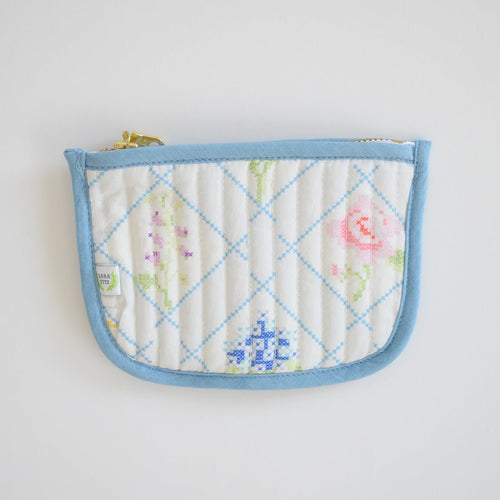 The Sara Fitz Coin Pouch, Cross Stitch Floral, is a quilted cotton card holder with a light blue border and zip closure, featuring pastel cross-stitched florals on white.