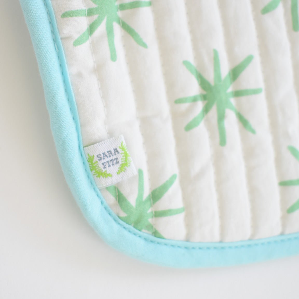 A close-up of the Sara Fitz Coin Pouch, Green Star shows a white cotton quilted bag with light green starburst patterns, a blue border, and a small fabric tag labeled Sara Fitz in green and blue.
