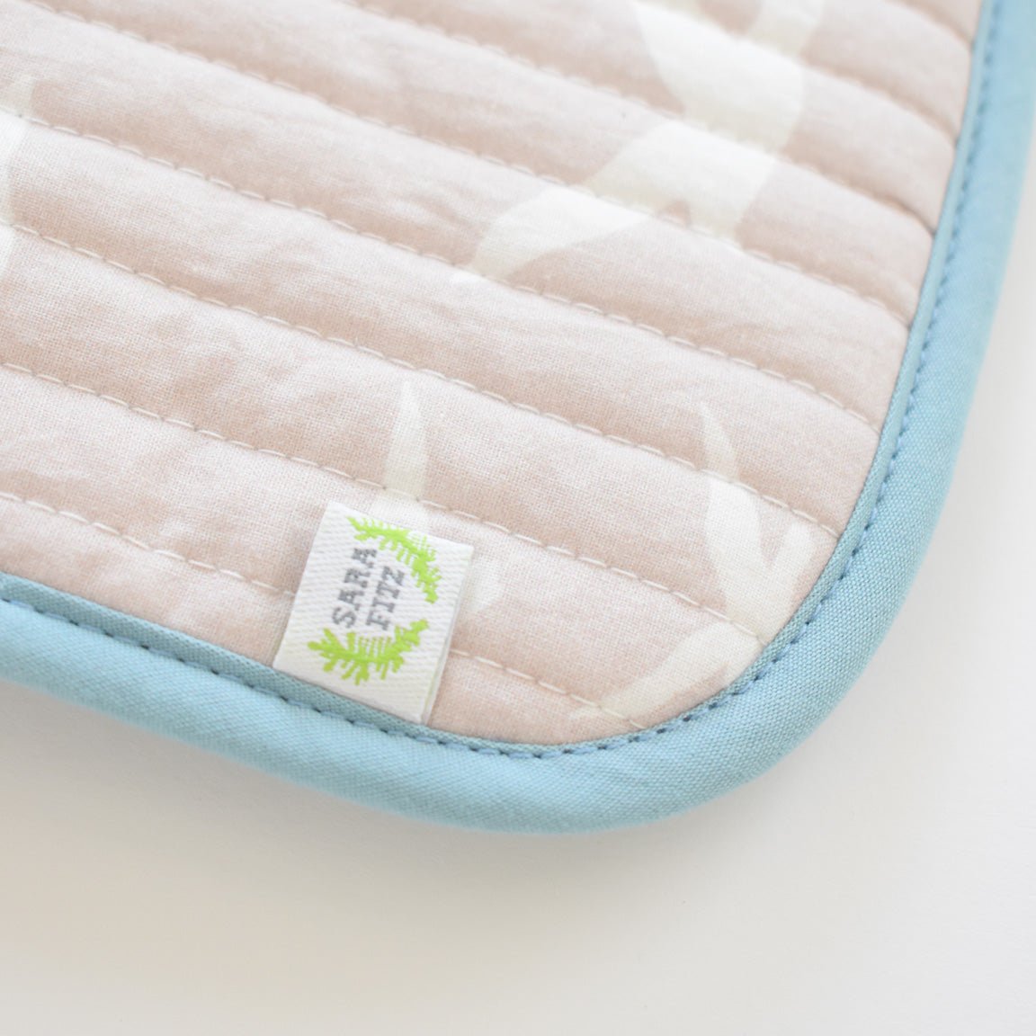 Close-up of the Sara Fitz Coin Pouch, Seagull—a quilted cotton card holder featuring a light pink and white pattern, blue edging, and a small white tag with green leaf detail. This handmade accessory from India rests on a white surface.