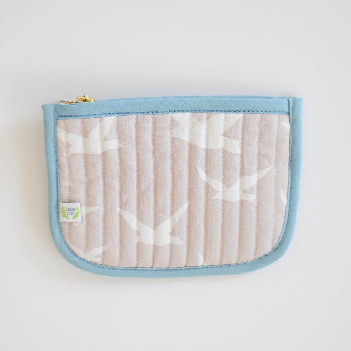 The Sara Fitz Coin Pouch, Seagull, features a beige fabric with a white bird pattern, light blue trim, a gold zipper at the top left corner, and a small green and white label. Inspired by handmade bag designs from India.