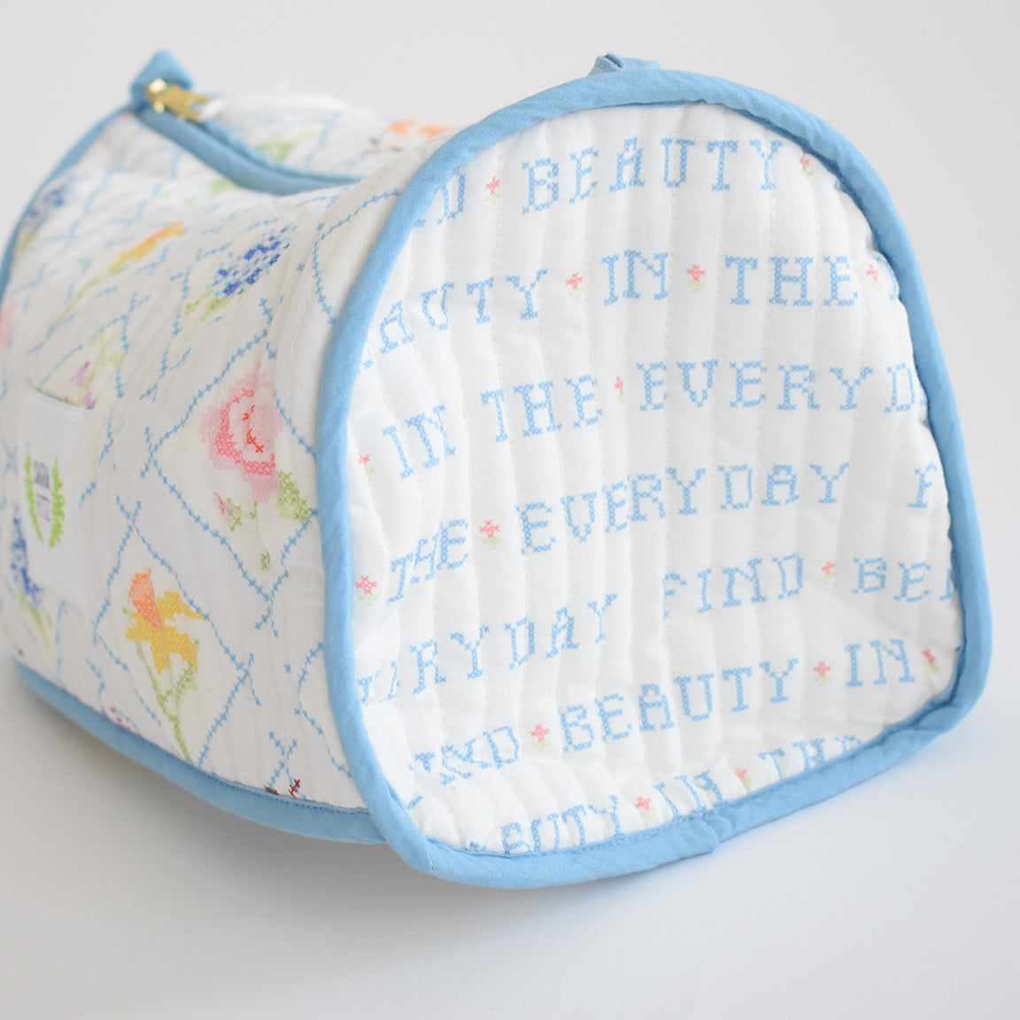 The Sara Fitz Cosmetic Pouch, Cross Stitch Floral, is a quilted cotton bag with blue trim and pastel cross-stitched florals. "FIND BEAUTY IN THE EVERYDAY" is stitched in blue on the side.