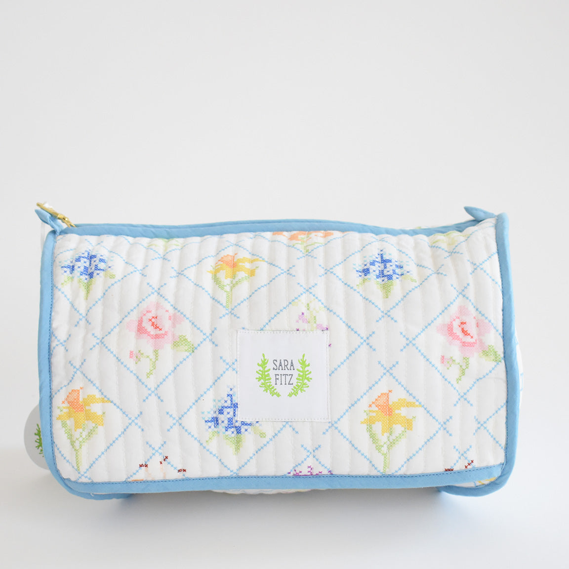 The Cosmetic Pouch, Cross Stitch Floral by Sara Fitz is a rectangular quilted cotton bag with light blue trim and a pastel floral pattern. The front has a white label with "Sara Fitz" in green. The background is plain white.
