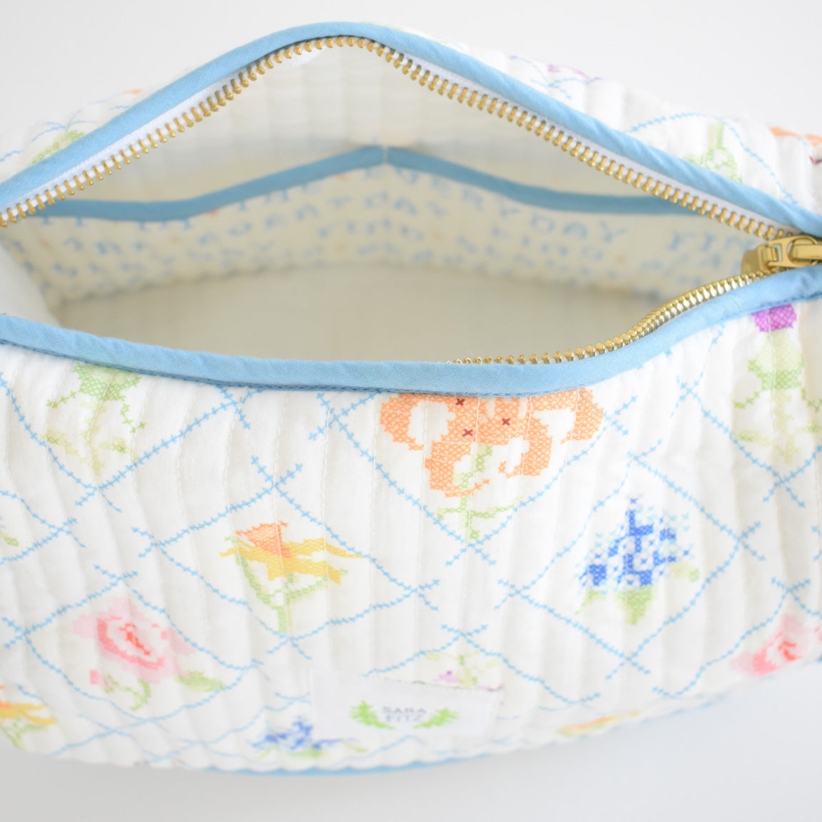 The Sara Fitz Cosmetic Pouch, Cross Stitch Floral, is a quilted cotton bag with a blue zipper, featuring a colorful floral and blue check pattern. Its light fabric lining is printed with small blue letters—ideal as a travel organizer or toiletry bag.