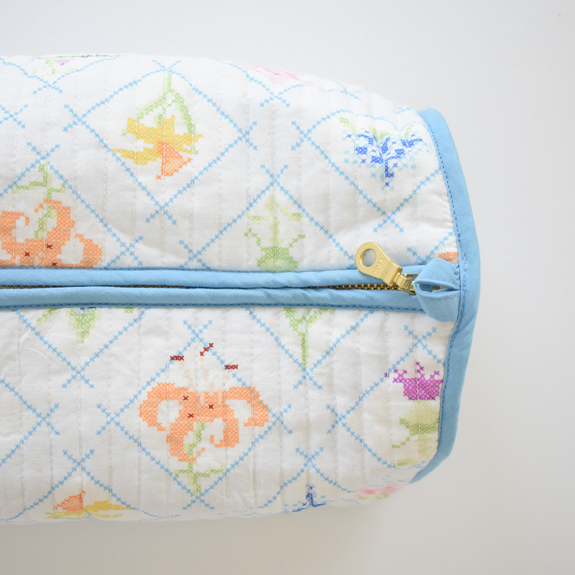 The Sara Fitz Cosmetic Pouch, Cross Stitch Floral, features a cylindrical quilted cotton design with a light blue zipper and trim, decorated in colorful pixelated florals within a blue diamond grid—ideal for use as a toiletry bag or travel organizer.