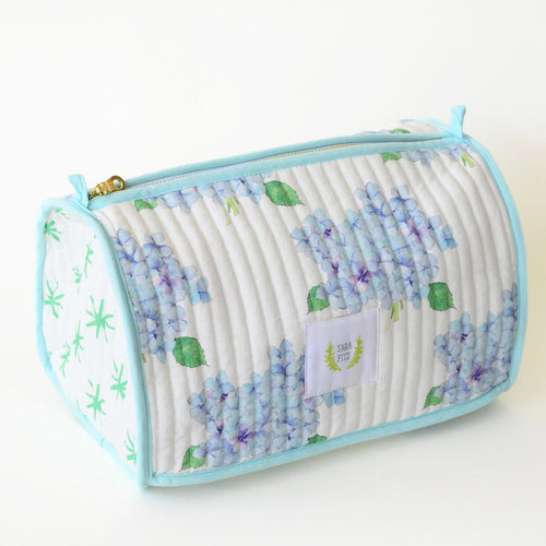 The Sara Fitz Cosmetic Pouch, Hydrangea, is a quilted cotton organizer with light blue trim, adorned with blue and purple hydrangeas and green leaves. It features a top zipper and a white front label.
