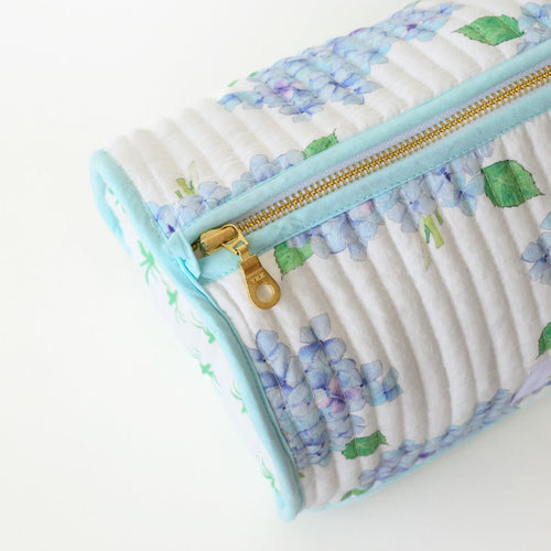 A close-up of the Sara Fitz Cosmetic Pouch, Hydrangea—a quilted cotton bag with a gold zipper, showcasing light blue and purple hydrangeas with green leaves on white, light blue trim, perfect as a travel organizer.