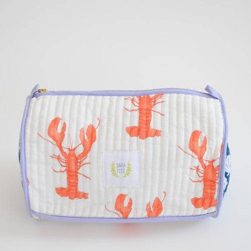 The Sara Fitz Cosmetic Pouch, Lobster, features orange lobster prints on a white quilted cotton bag with light purple trim. Ideal for toiletries or travel essentials, it has a small center label reading “Sara Fitz.”.