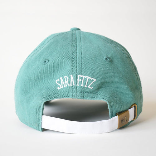 A moss green cotton baseball cap by Sara Fitz, shown from the back, features "SARA FITZ" in white needlepoint embroidery above an adjustable white strap with a brass buckle engraved with "HL.