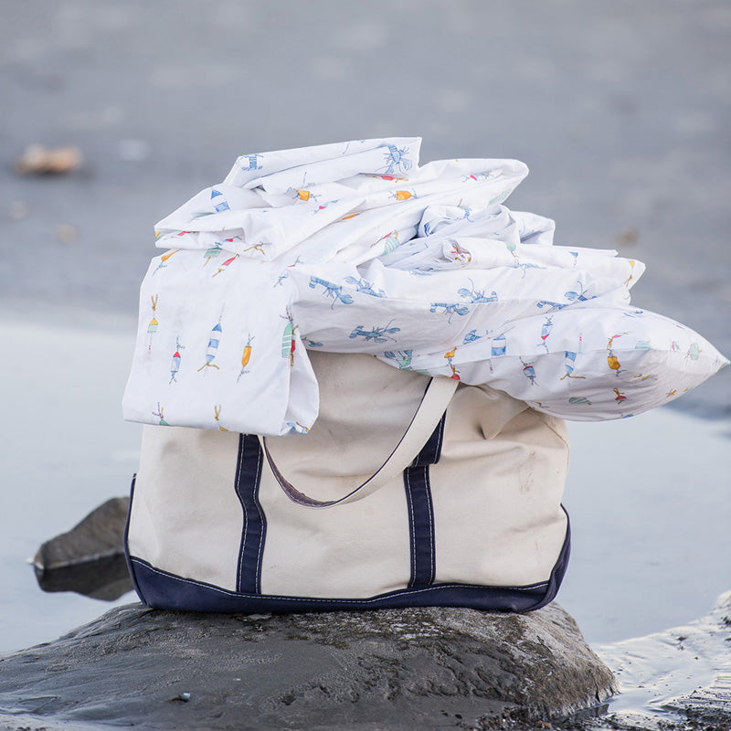 A white tote bag with navy blue trim sits on a rock by the water, topped with a loosely folded, white patterned sheet or blanket. The shoreline and wet sand are visible in the background.