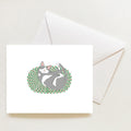 A Cozy Kitty Note Card features a gray and white cat curled up sleeping on a green and pink braided rug, displayed next to an envelope.