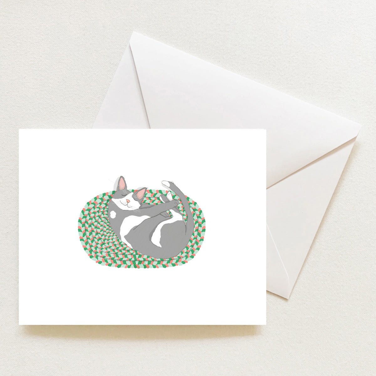 A Cozy Kitty Note Card features a gray and white cat curled up sleeping on a green and pink braided rug, displayed next to an envelope.