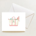 A Holiday Tote Note Card from Sara Fitz featuring a watercolor illustration of a canvas tote bag filled with holiday items, including candy canes and berries, shown with a matching envelope.