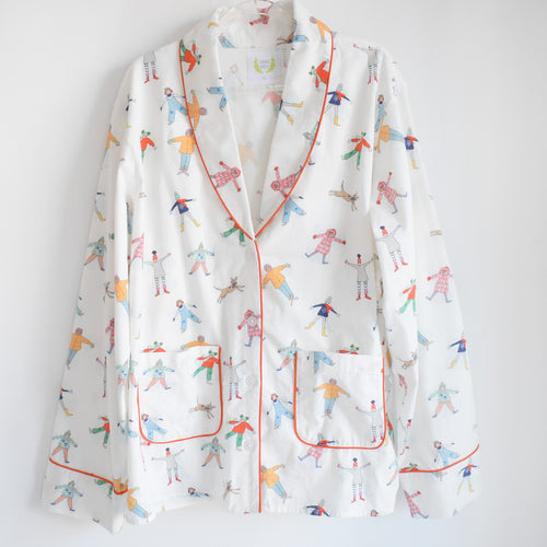 The Snow Angels Pajama Set by Sara Fitz features a white brushed cotton long sleeve top with colorful winter figures and orange piping, paired with matching wide-leg bottoms. Set is shown on a plain white background.