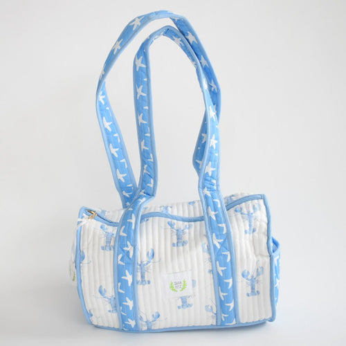 The Sara Fitz Purse, Blue Lobster, is a handmade white cotton shoulder bag with light blue trim, rectangular shape, two long straps, and a pattern of blue lobsters and white birds. Made in India.