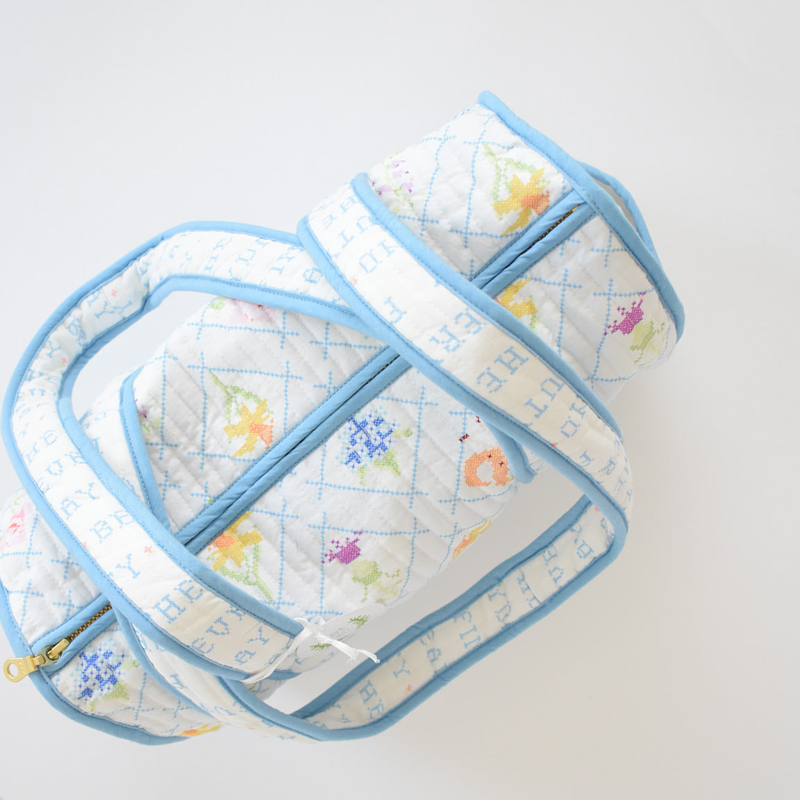 The Sara Fitz Purse, Blue Cross Stitch Floral, is a handmade cotton shoulder bag from India featuring a quilted texture, light blue trim and handles, visible zipper, and charming floral cross stitch on a white background.