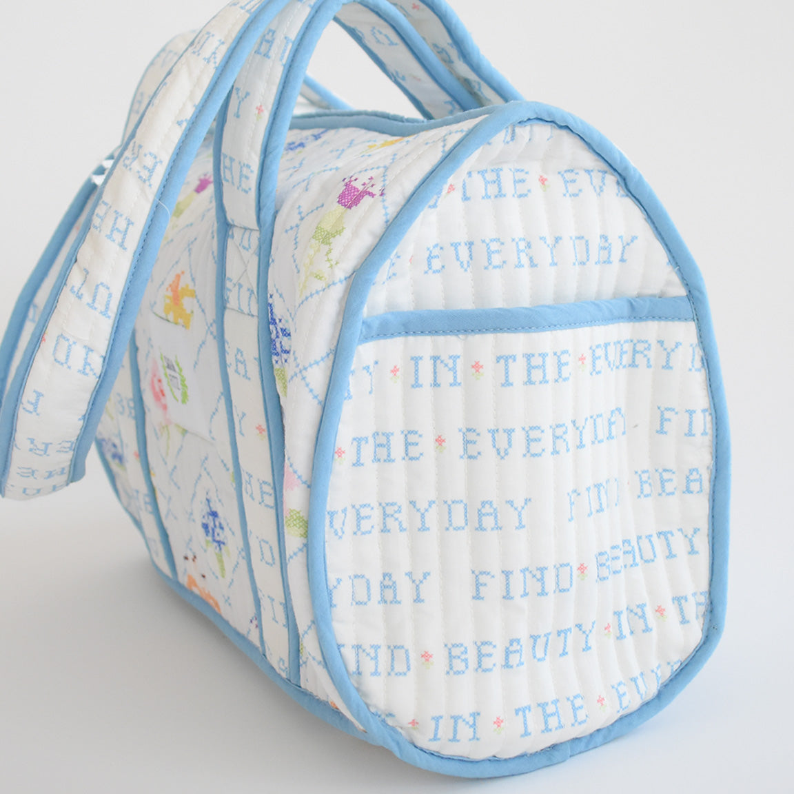 Handmade in India, the Sara Fitz Purse features a white quilted cotton design with light blue trim and handles, blue "FIND BEAUTY IN THE EVERYDAY" text, and small pastel cross stitch floral accents.