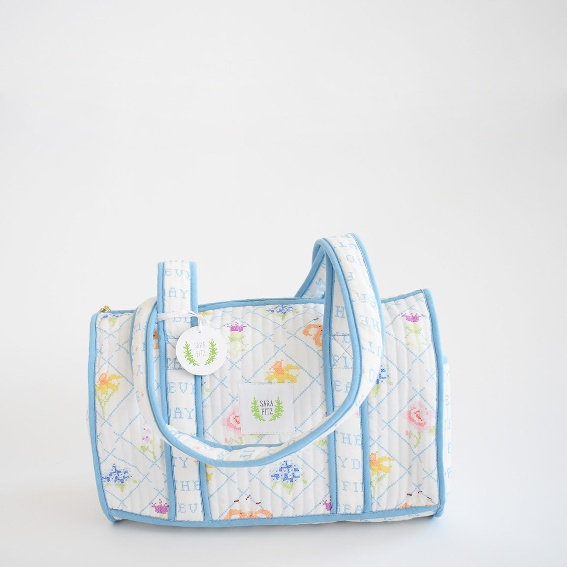 The Sara Fitz Purse, Blue Cross Stitch Floral, is a cotton shoulder bag with pastel floral patterns, blue trim and handles. Handmade in India, it features a round tag with a green tree and HEYDAY in a light blue grid pattern.