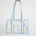 The Sara Fitz Purse, Blue Cross Stitch Floral, is a quilted cotton shoulder bag with pale blue trim and handles, featuring a pastel floral and letter design, zip closure, and a small round tag on one handle.