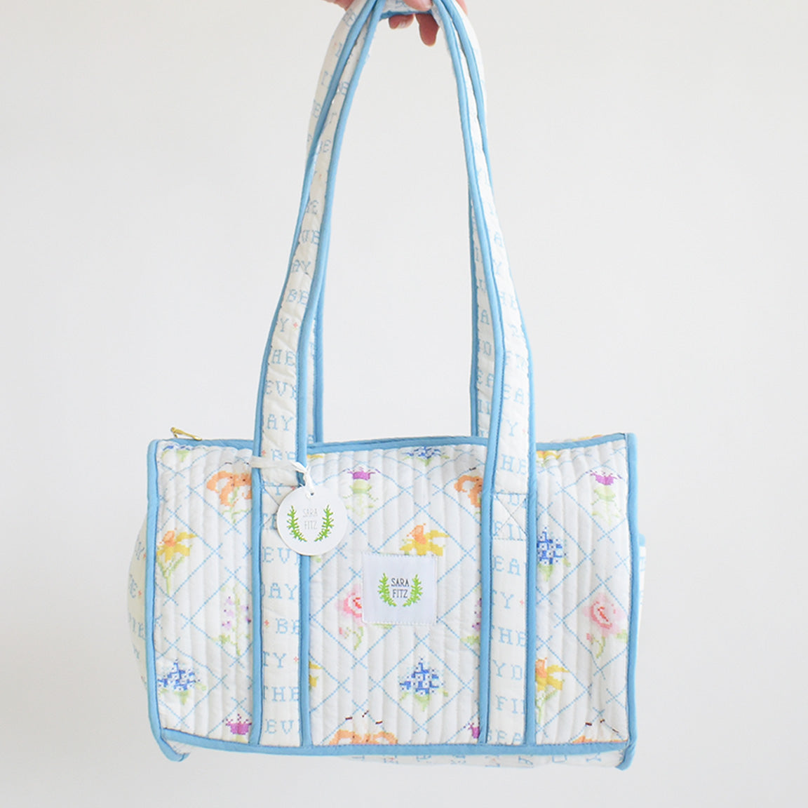 The Sara Fitz Purse, Blue Cross Stitch Floral, is a quilted cotton shoulder bag with pale blue trim and handles, featuring a pastel floral and letter design, zip closure, and a small round tag on one handle.