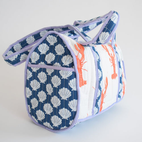 The Sara Fitz Purse, Lobster is a handmade quilted shoulder bag from India, with blue and white shell patterns on the sides, orange lobster prints in the center, and light purple piping and handles.